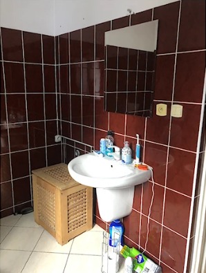 Łazienka z brązowymi płytkami, białą umywalką na postumencie, drewnianą szafką z lamelami i prostokątnym lustrem na ścianie.