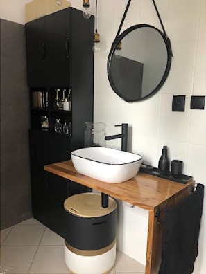 Nowoczesna łazienka z białą umywalką nablatową z czarnym obramowaniem, drewnianym blatem i okrągłym lustrem na skórzanym pasku, czarna szafka z otwartymi półkami na kosmetyki.
