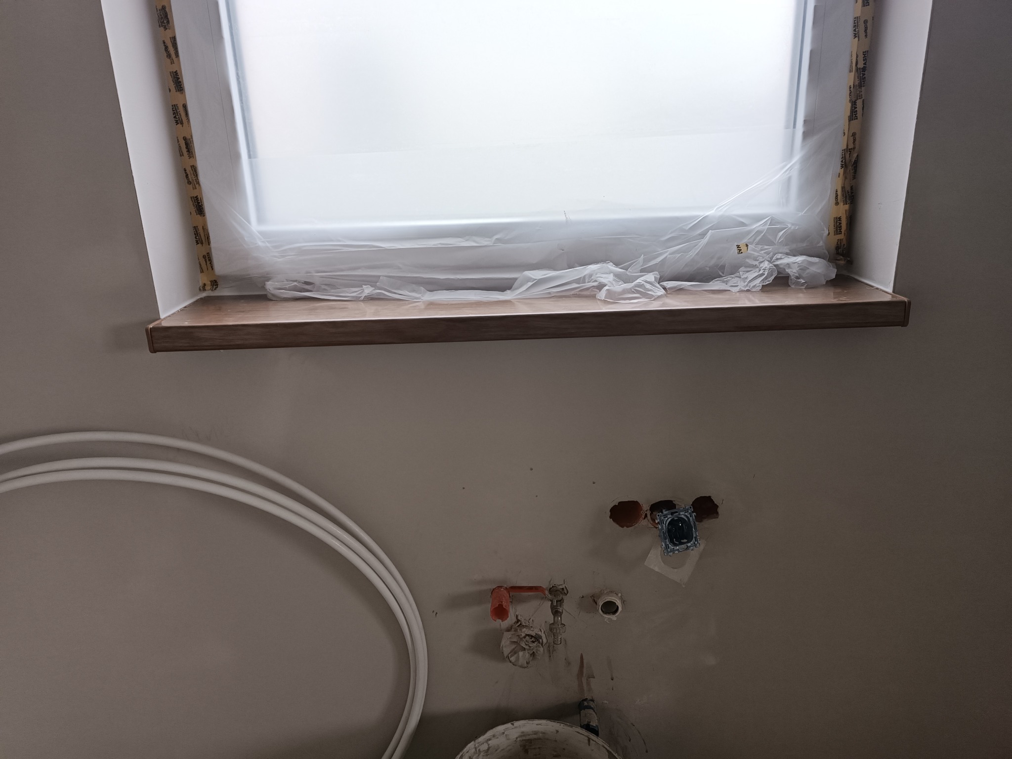 Montaż drewnianego parapetu okiennego w trakcie prac wykończeniowych, okno zabezpieczone folią, widoczne instalacje wodne i elektryczne w ścianie.