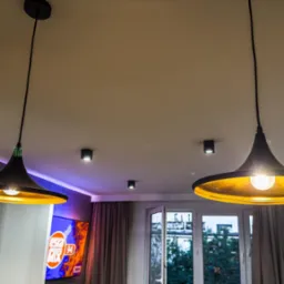 Dwa czarne, stożkowe klosze lamp wiszących z żółtym wnętrzem, na tle sufitu z trzema małymi lampami punktowymi i oknem balkonowym z widokiem na drzewa.