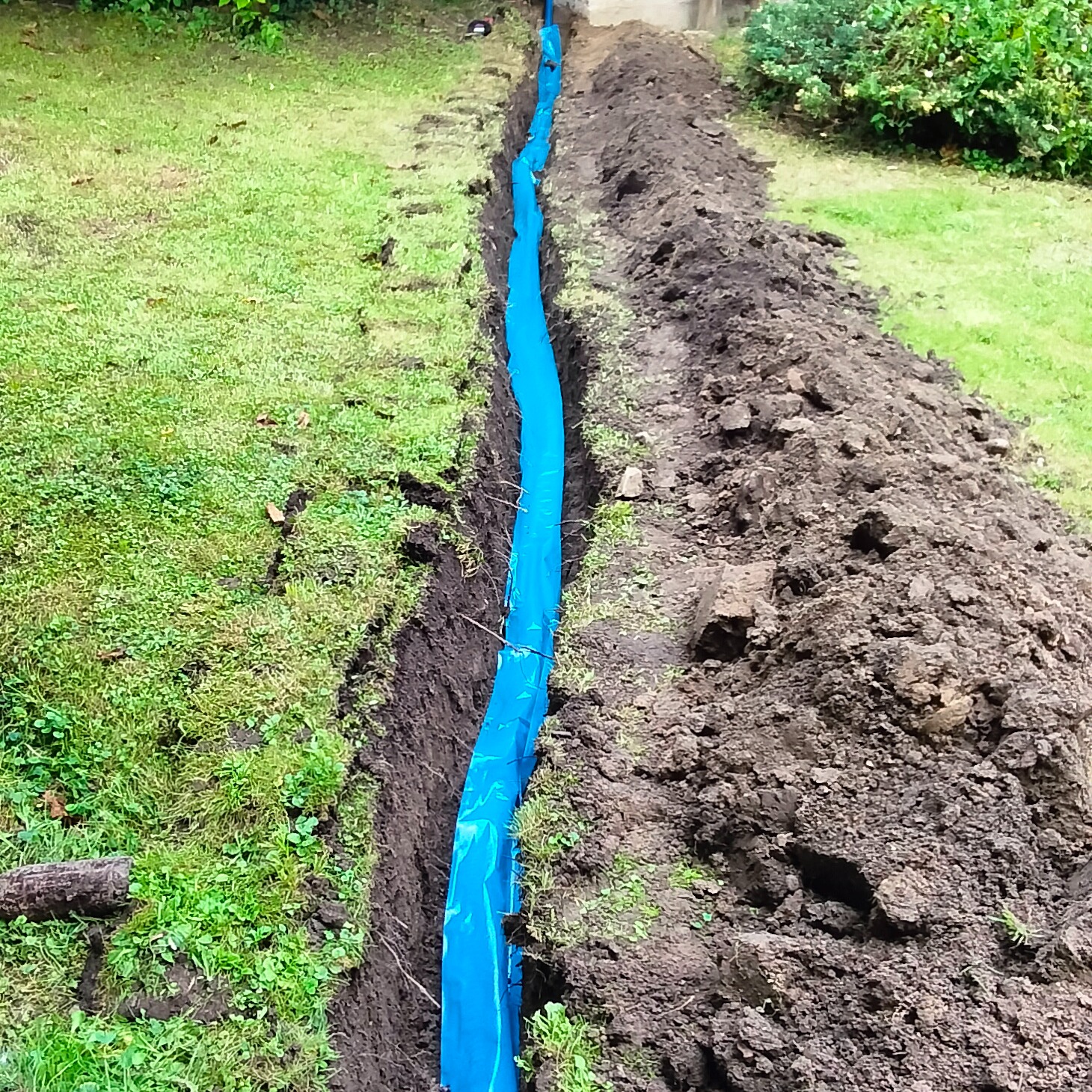 Wykop w ziemi, zabezpieczony niebieską folią, prawdopodobnie pod kable elektryczne, widok z góry na trawnik i pryzmę ziemi.