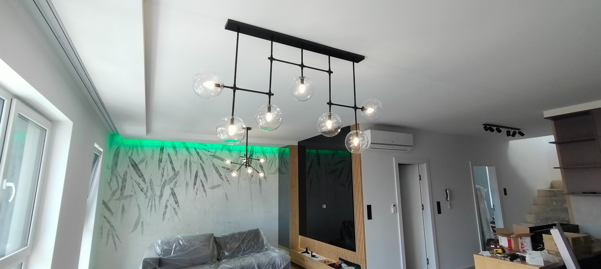 Nowoczesny salon z zamontowanym oświetleniem: czarny żyrandol z kulistymi kloszami, dodatkowe oświetlenie punktowe na suficie i podświetlenie LED za kanapą.