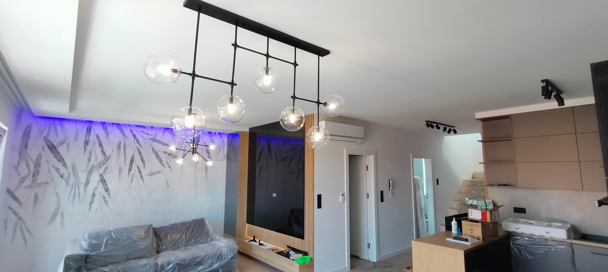 Nowoczesny salon z zainstalowanym czarnym, metalowym żyrandolem z kulistymi szklanymi kloszami, widoczny również drugi żyrandol, ściana z motywem roślinnym podświetlona fioletowym światłem LED.