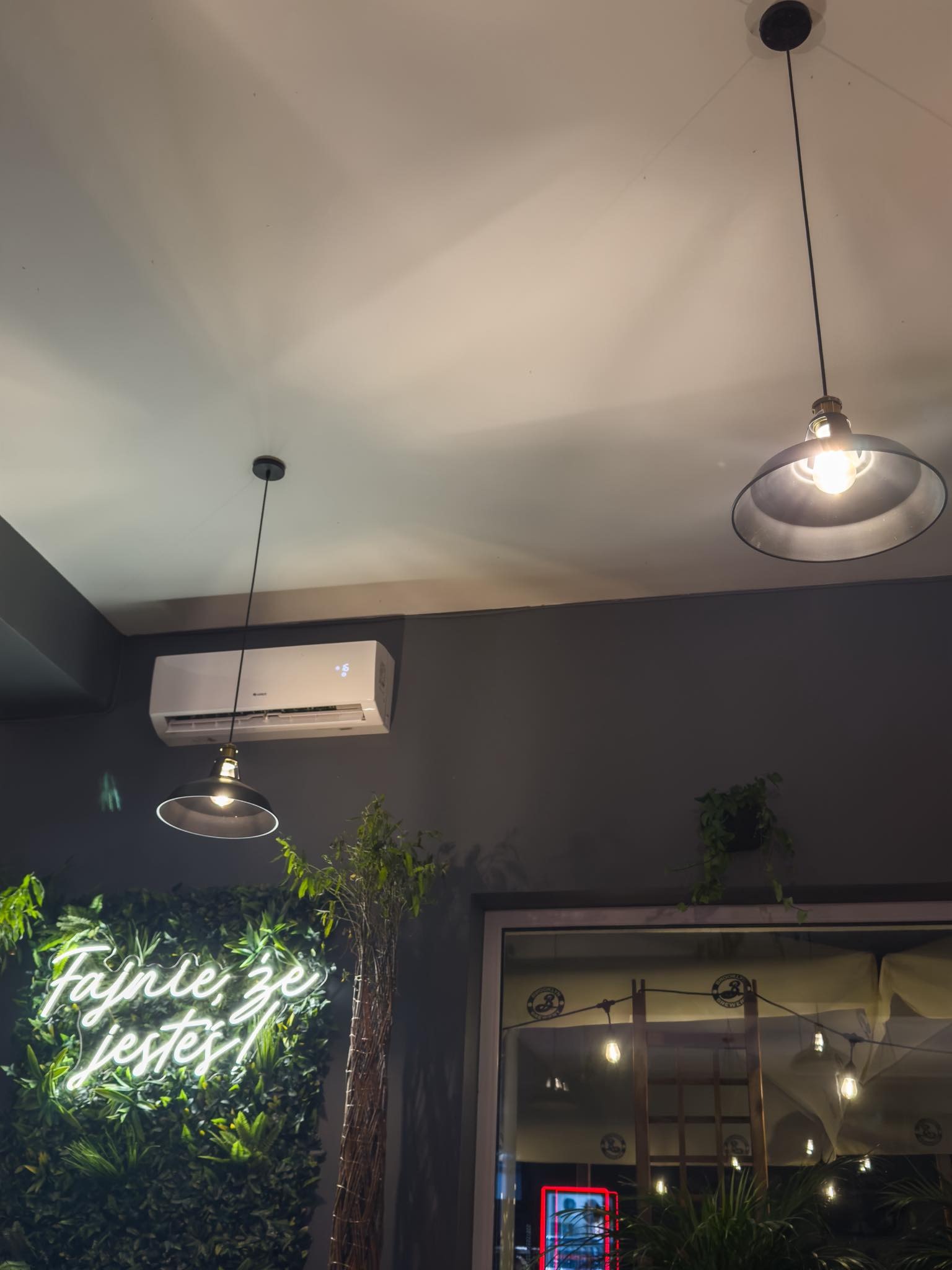 Dwa metalowe klosze lamp wiszących na tle sufitu i szarej ściany z klimatyzatorem, neonem 'Fajnie, że jesteś!' na tle roślinnym i oknem z dekoracyjnym oświetleniem.