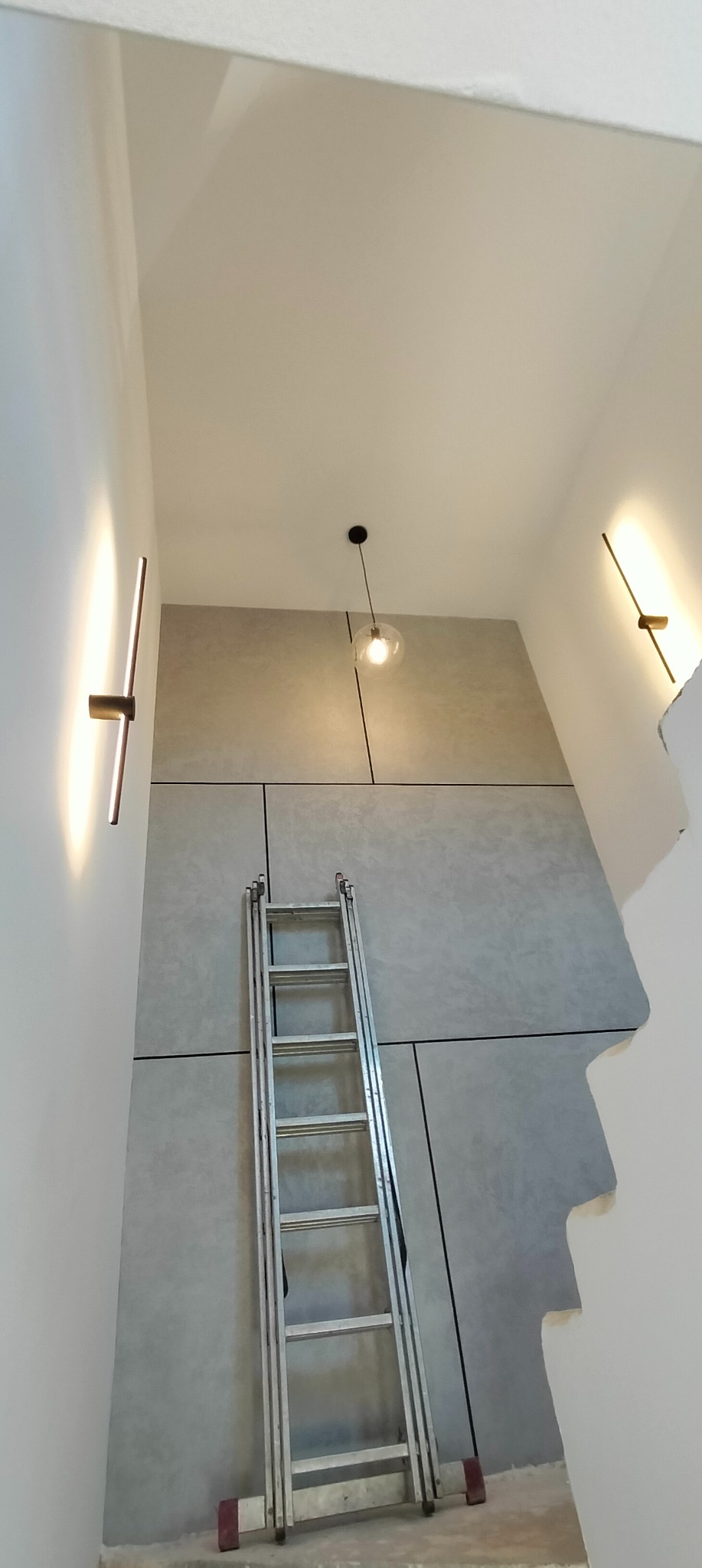 Wysokie wnętrze z szarą ścianą wykończoną panelami, oświetlone lampami ściennymi i wiszącą lampą kulą, z rozłożoną aluminiową drabiną opartą o ścianę.