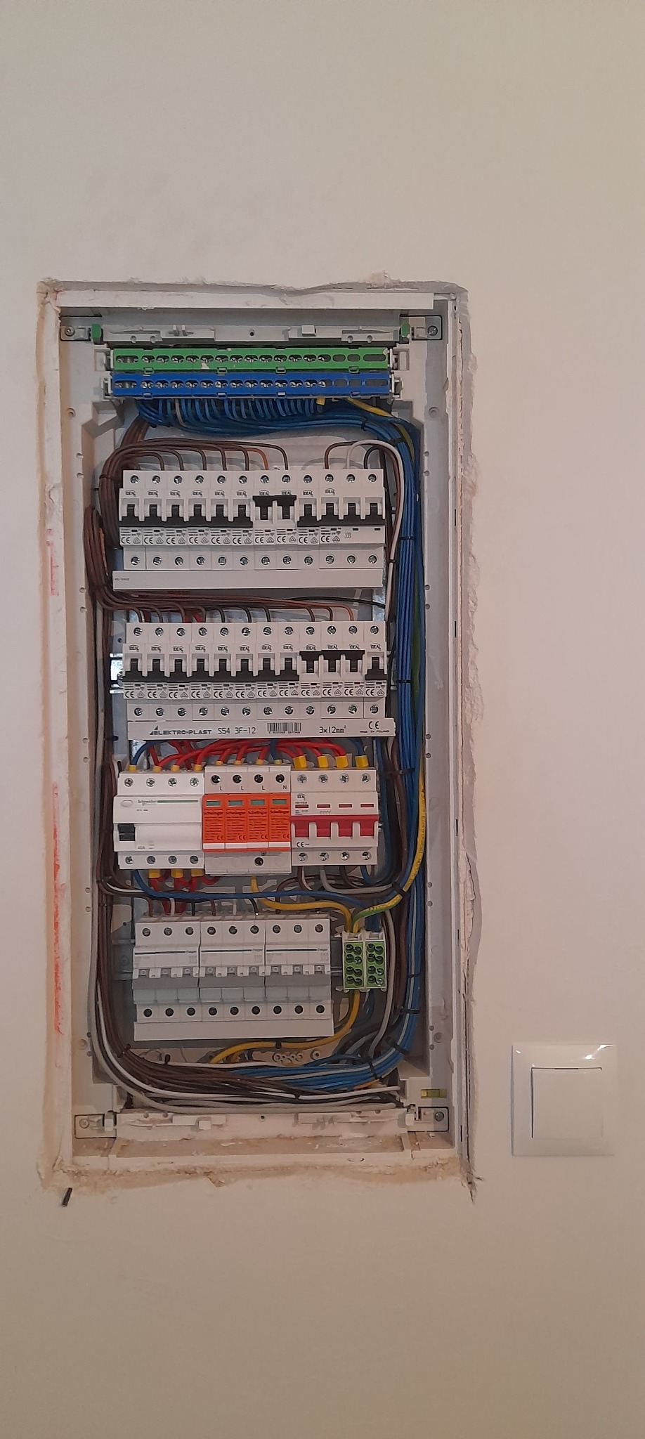Rozdzielnica elektryczna z zainstalowanymi bezpiecznikami, okablowaniem i listwami zaciskowymi, widoczne ślady po montażu w ścianie, obok włącznik światła.