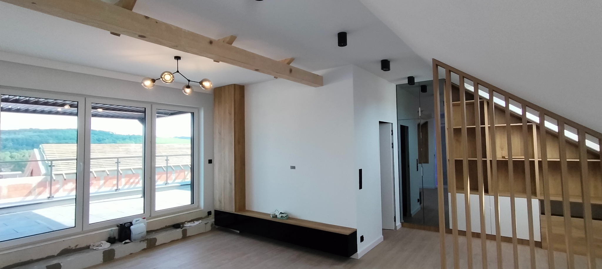 Jasne poddasze z drewnianymi belkami, nowoczesnym oświetleniem sufitowym i dużymi oknami balkonowymi z widokiem na zieleń. Widoczne czarne punkty oświetleniowe na suficie, drewniana zabudowa ściany...