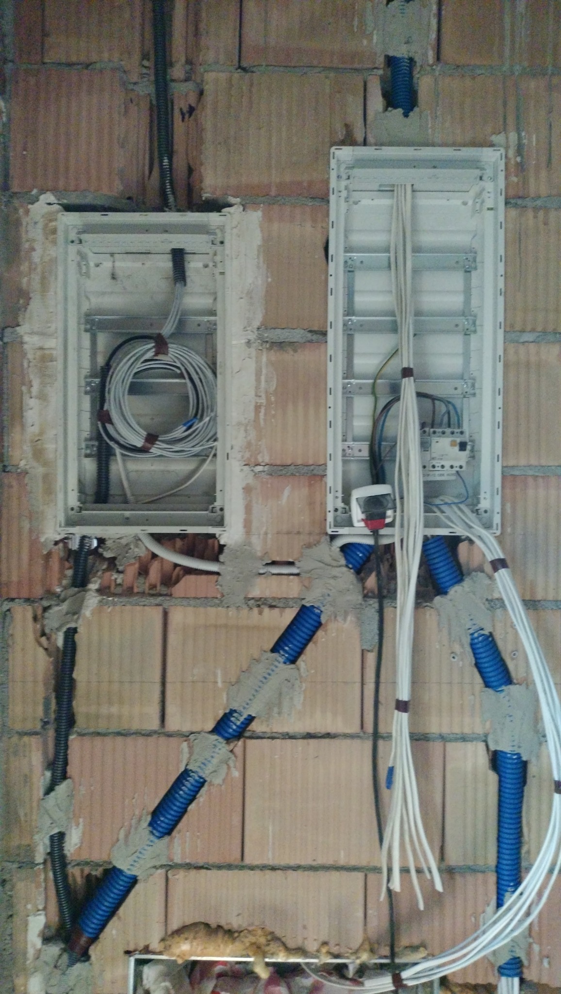 Dwa metalowe panele elektryczne zamontowane w surowej ceglanej ścianie, z widocznymi niebieskimi rurkami i przewodami elektrycznymi zabezpieczonymi zaprawą.
