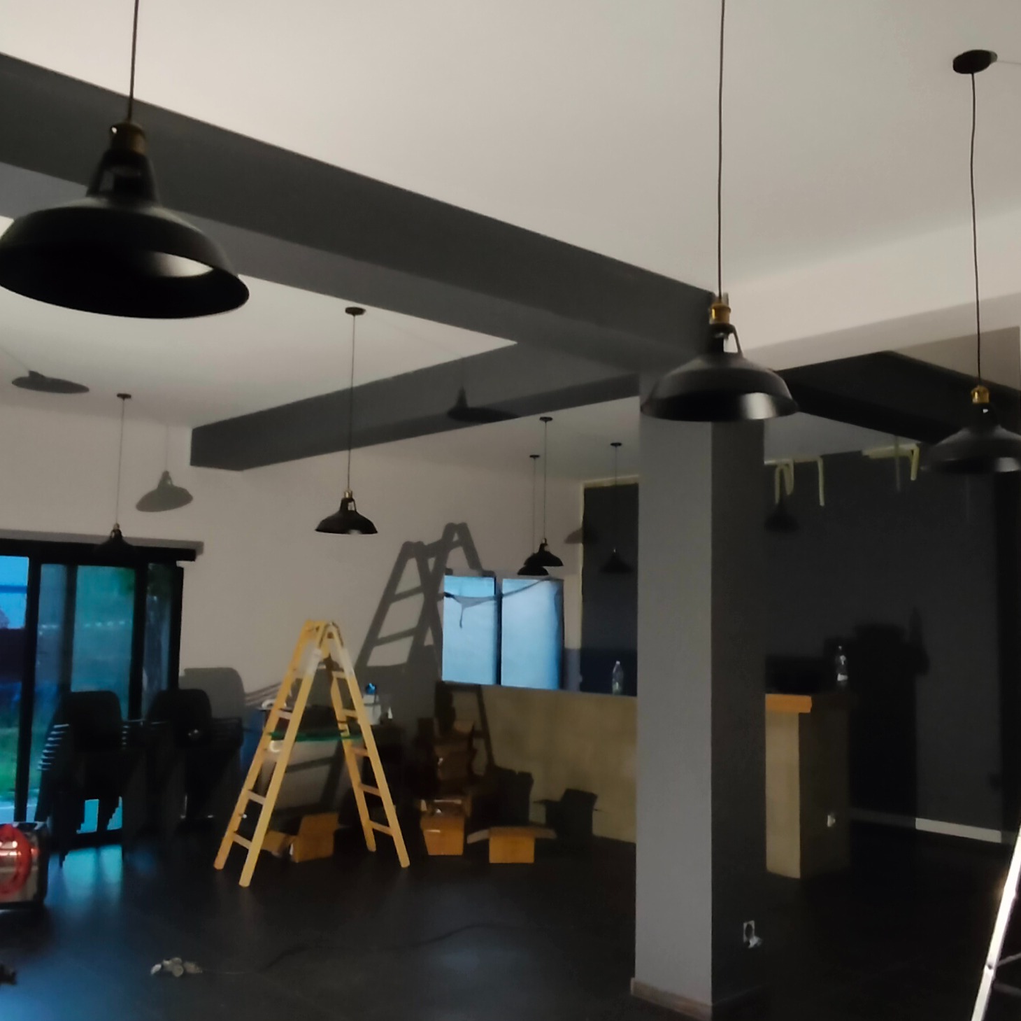 Wnętrze lokalu w trakcie montażu oświetlenia: czarne, metalowe lampy wiszące z żółtymi elementami, widoczne drabiny i przygotowane podłoże, surowy wystrój.