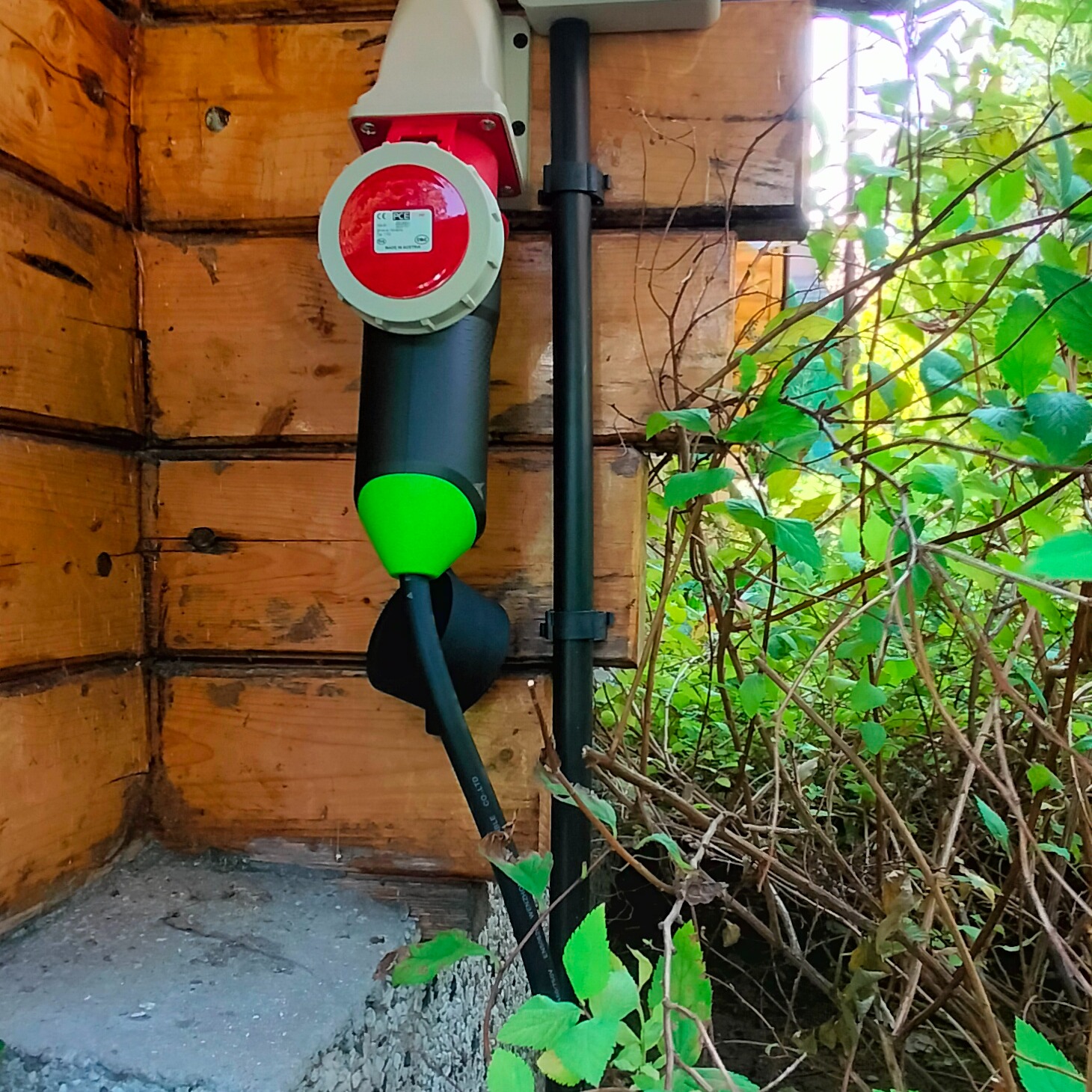 Zewnętrzne gniazdo siłowe, czerwono-białe, z podłączonym kablem zakończonym zieloną wtyczką, zamontowane na drewnianej ścianie budynku, z widocznymi roślinami w tle.