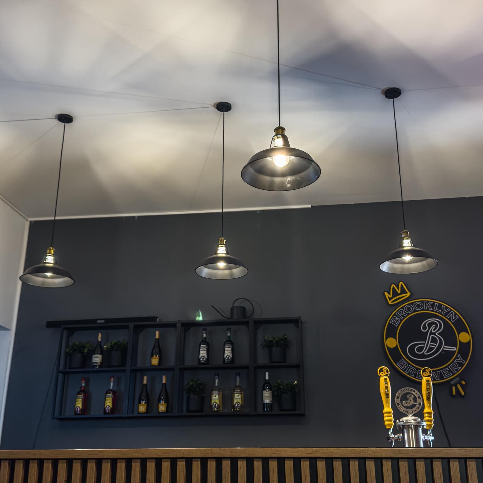 Cztery metalowe lampy wiszące z żarówkami Edison nad barem z ciemnoszarą ścianą, półkami z butelkami i logo Brooklyn Brewery.