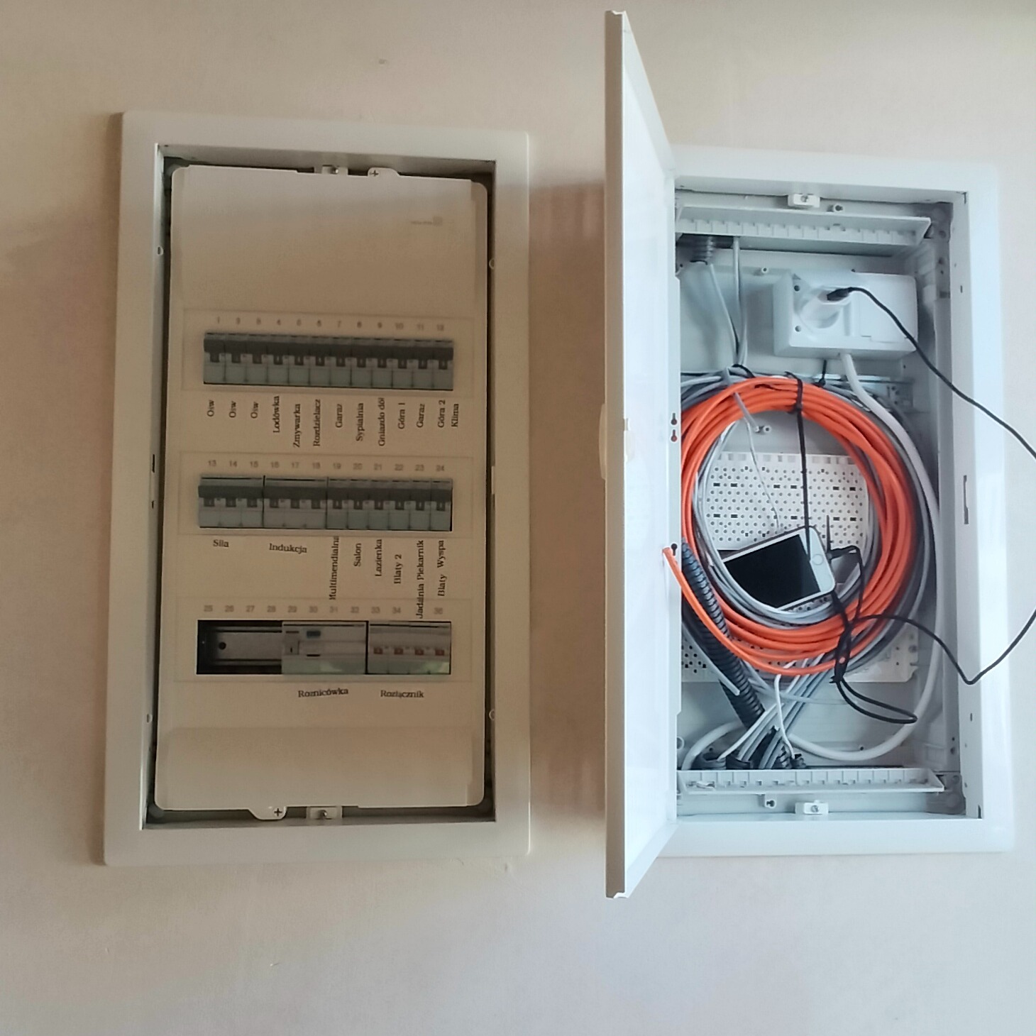 Otwarta skrzynka rozdzielcza z widocznymi kablami, w tym pomarańczowy przewód zwinięty w okrąg, oraz smartfon wewnątrz, obok zamknięta skrzynka z opisem obwodów.