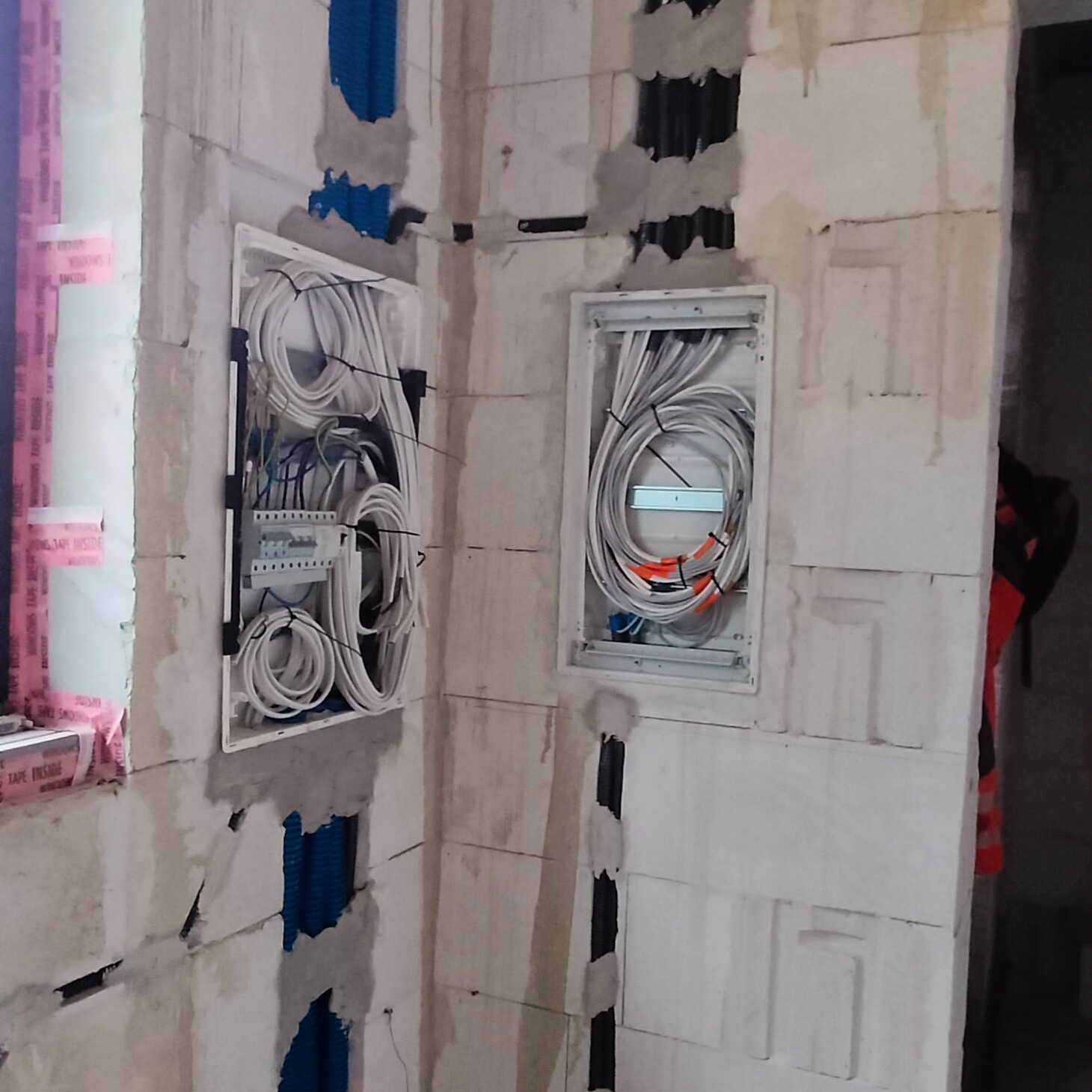 Dwie podtynkowe skrzynki elektryczne z przewodami w ścianie z betonu komórkowego, widoczne niebieskie rury karbowane i czarne peszle, ściany w stanie surowym.