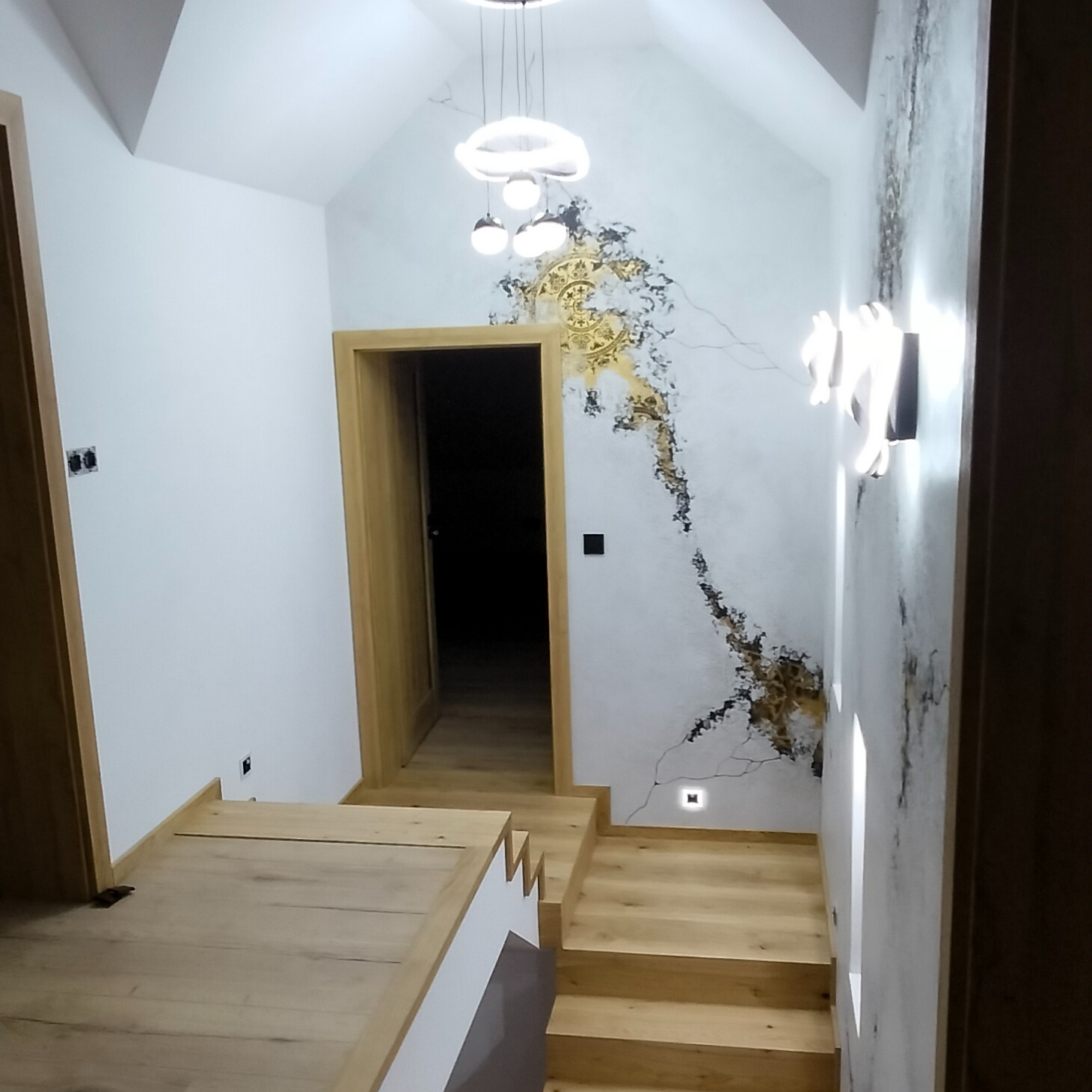 Nowoczesne oświetlenie schodów: wisząca lampa z kloszami w kształcie pierścieni i kinkiet ścienny z podświetleniem LED, dekoracyjna ściana z motywem złotych pęknięć, drewniane schody...