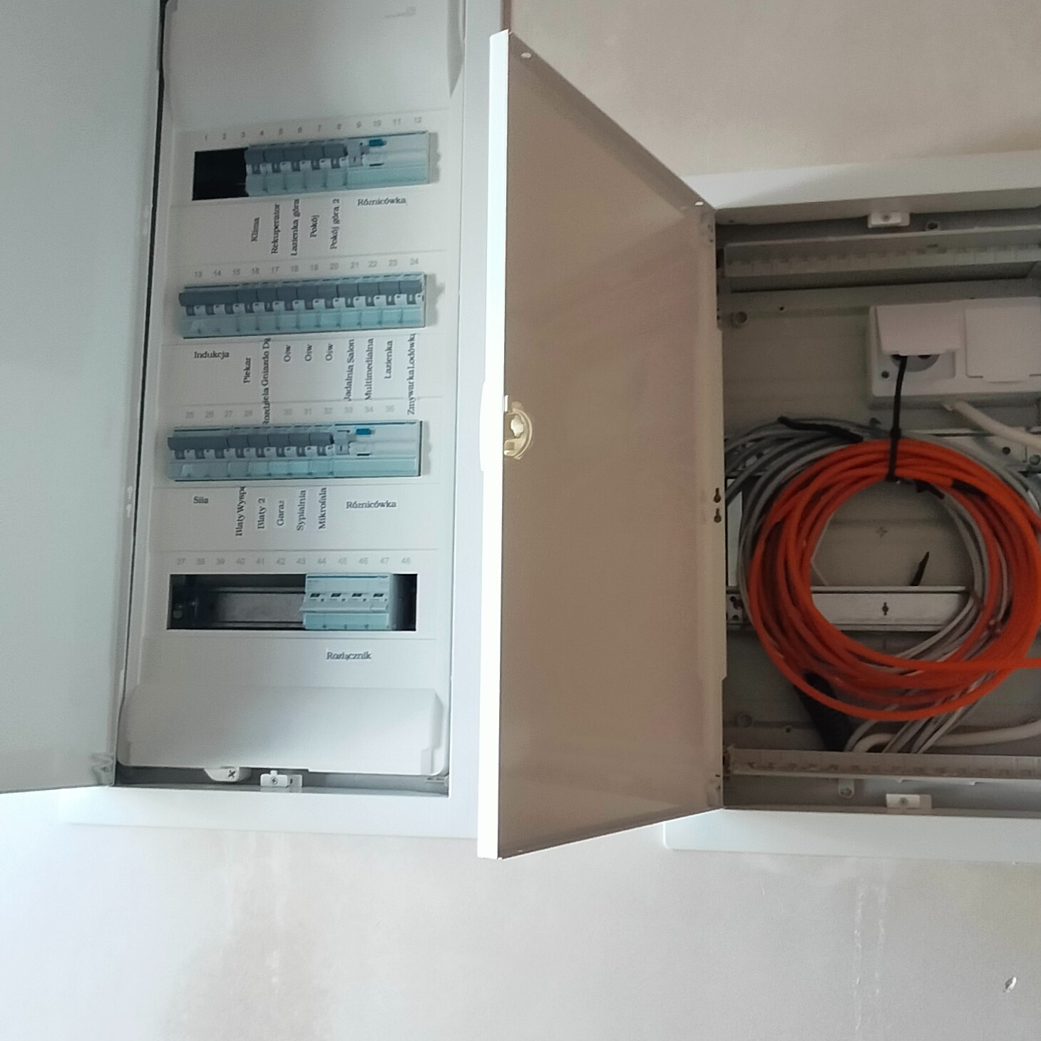 Otwarta skrzynka rozdzielcza z widocznymi bezpiecznikami, opisami obwodów i zwiniętym pomarańczowym przewodem wewnątrz, ukazująca etap prac instalacyjnych.
