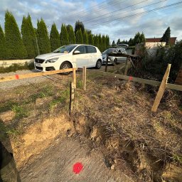 Bernadeta Kargul - Wytyczanie fundamentów pod budowę domu z użyciem drewnianych słupków i desek, widoczne oznaczenia geodezyjne w postaci czerwonych punktów na ziemi, w tle dwa samochody osobowe i rząd wysokich tui.