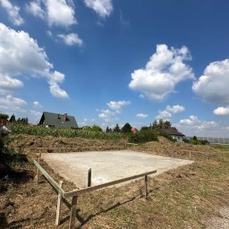 Bernadeta Kargul - Wylana betonowa płyta fundamentowa z drewnianym szalunkiem na działce z widokiem na domy i błękitne niebo z chmurami.