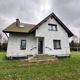 Bernadeta Kargul - Dwukondygnacyjny dom z szarą elewacją, brązowym dachem z zielonkawym nalotem i małym oknem dachowym, widoczny z perspektywy trawnika w pochmurny dzień.
