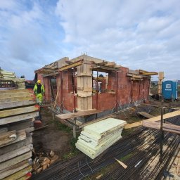 Bernadeta Kargul - Budowa domu jednorodzinnego w trakcie: widoczne ściany z czerwonej cegły, drewniane szalunki, izolacja termiczna, pracownik w odblaskowej kamizelce i przenośna toaleta TOI TOI w tle.
