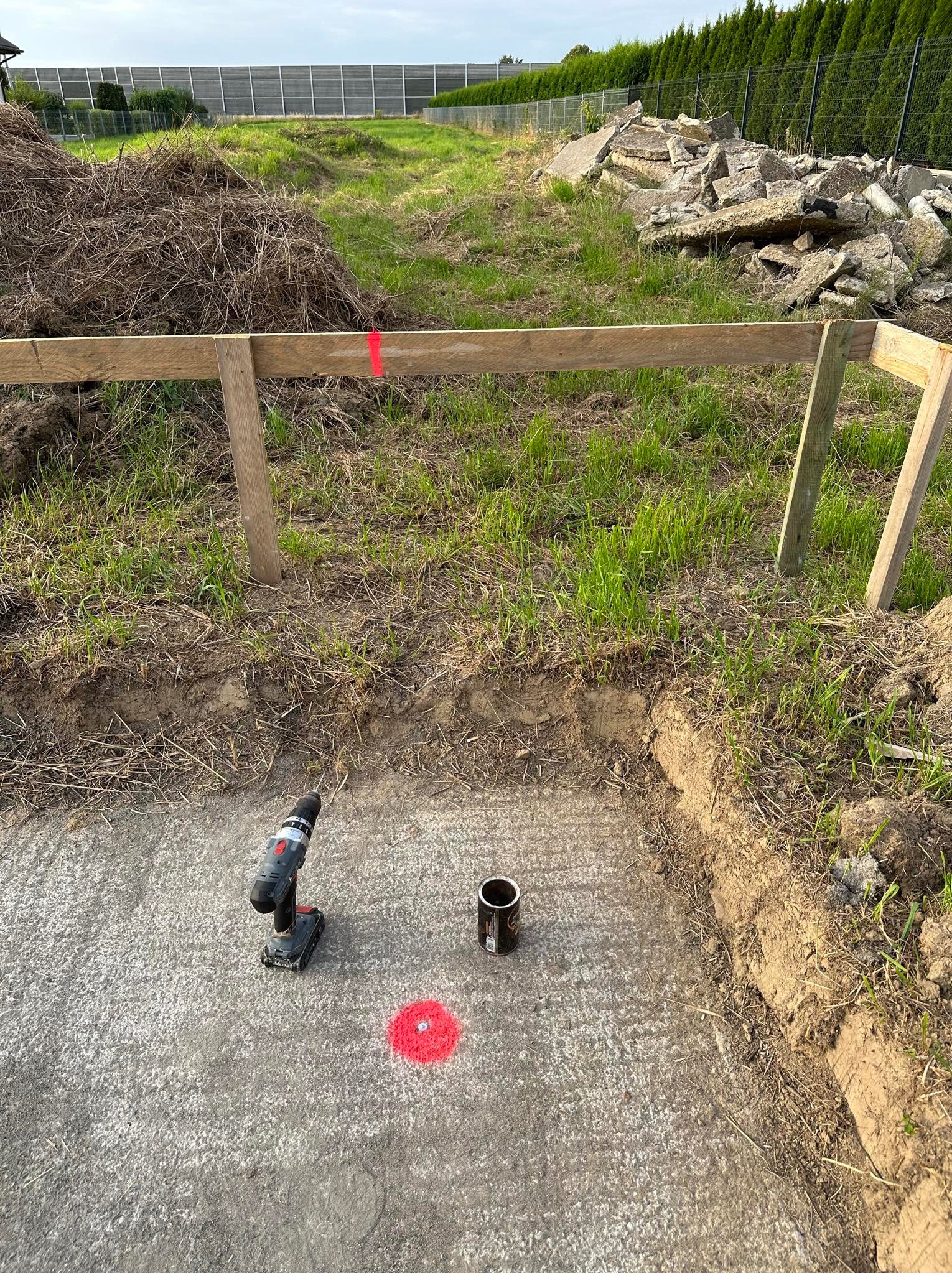 Przygotowanie fundamentu pod budowę: wykopany kwadrat z betonową wylewką, oznaczony czerwoną farbą, z wiertarką akumulatorową i puszką po napoju obok, drewniana konstrukcja szalunku z czerwoną...