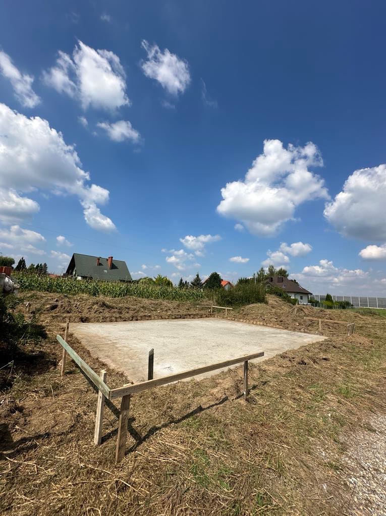 Wylana betonowa płyta fundamentowa z drewnianym szalunkiem na działce z widokiem na domy i błękitne niebo z chmurami.