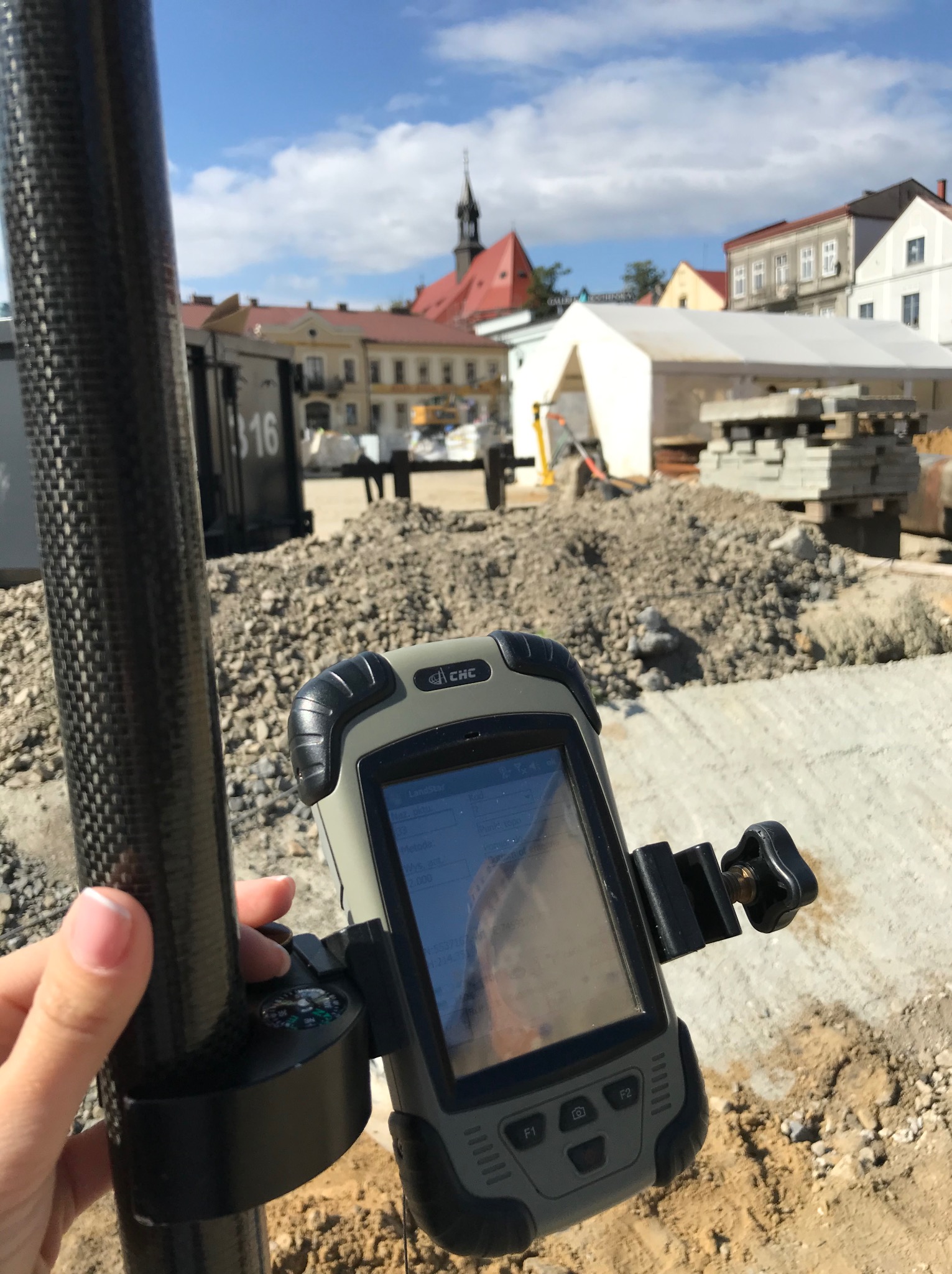 Geodezyjny odbiornik GPS marki CHC zamocowany na tyczce z kompasem, w tle plac budowy z gruzem, namiotem i budynkami w oddali pod błękitnym niebem.