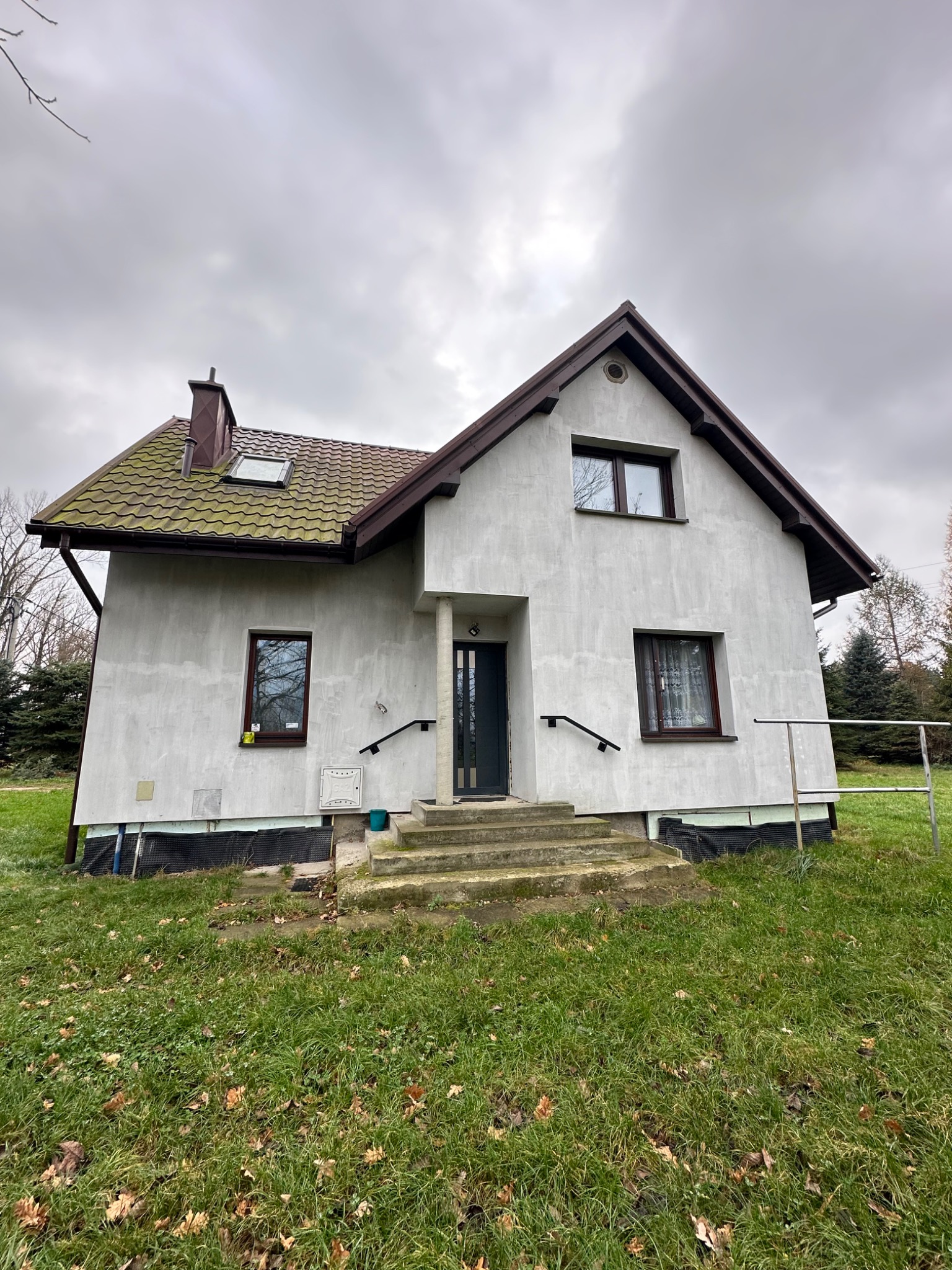 Dwukondygnacyjny dom z szarą elewacją, brązowym dachem z zielonkawym nalotem i małym oknem dachowym, widoczny z perspektywy trawnika w pochmurny dzień.