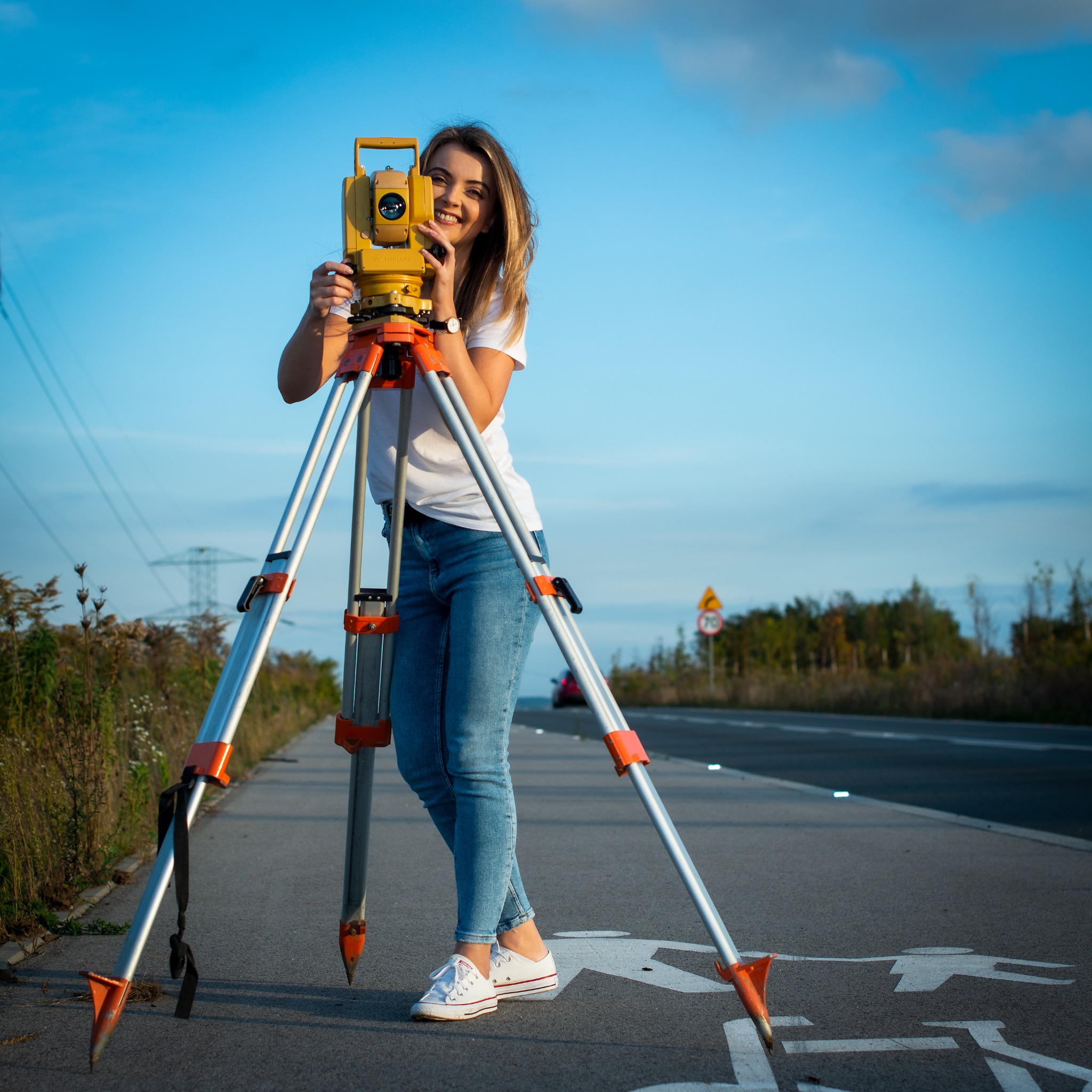 Uśmiechnięta geodetka w białej koszulce i jeansach obsługuje żółty teodolit Topcon na trójnogu, stojąc na ścieżce rowerowej obok drogi z widocznym znakiem ograniczenia prędkości do 70 km/h...