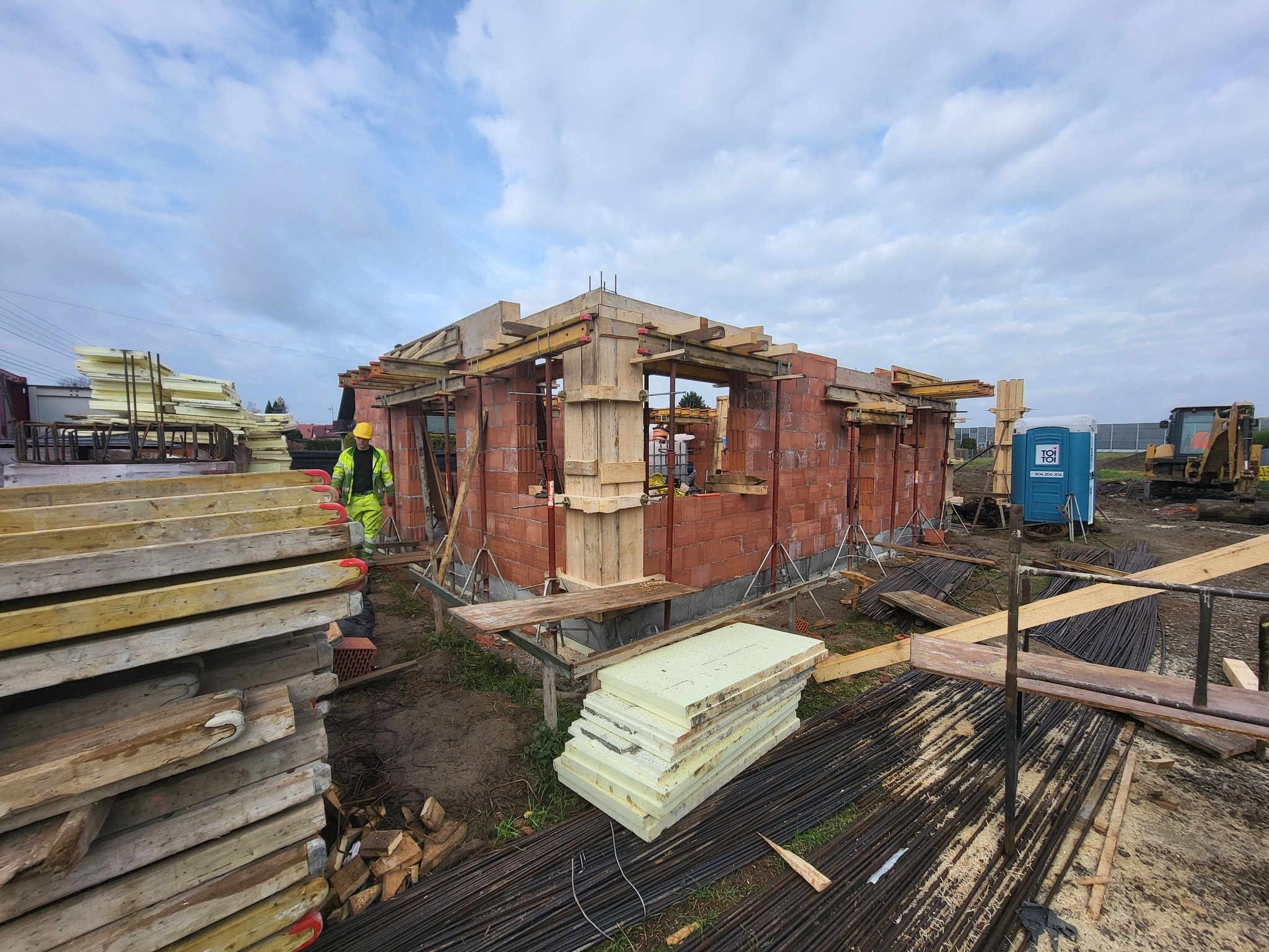 Budowa domu jednorodzinnego w trakcie: widoczne ściany z czerwonej cegły, drewniane szalunki, izolacja termiczna, pracownik w odblaskowej kamizelce i przenośna toaleta TOI TOI w tle.