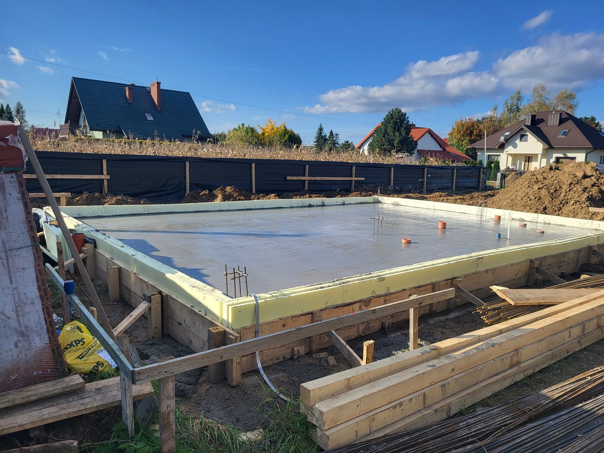 Wylana płyta fundamentowa z izolacją termiczną XPS, widoczne szalunki drewniane i wystające pręty zbrojeniowe, w tle zabudowa jednorodzinna i błękitne niebo.