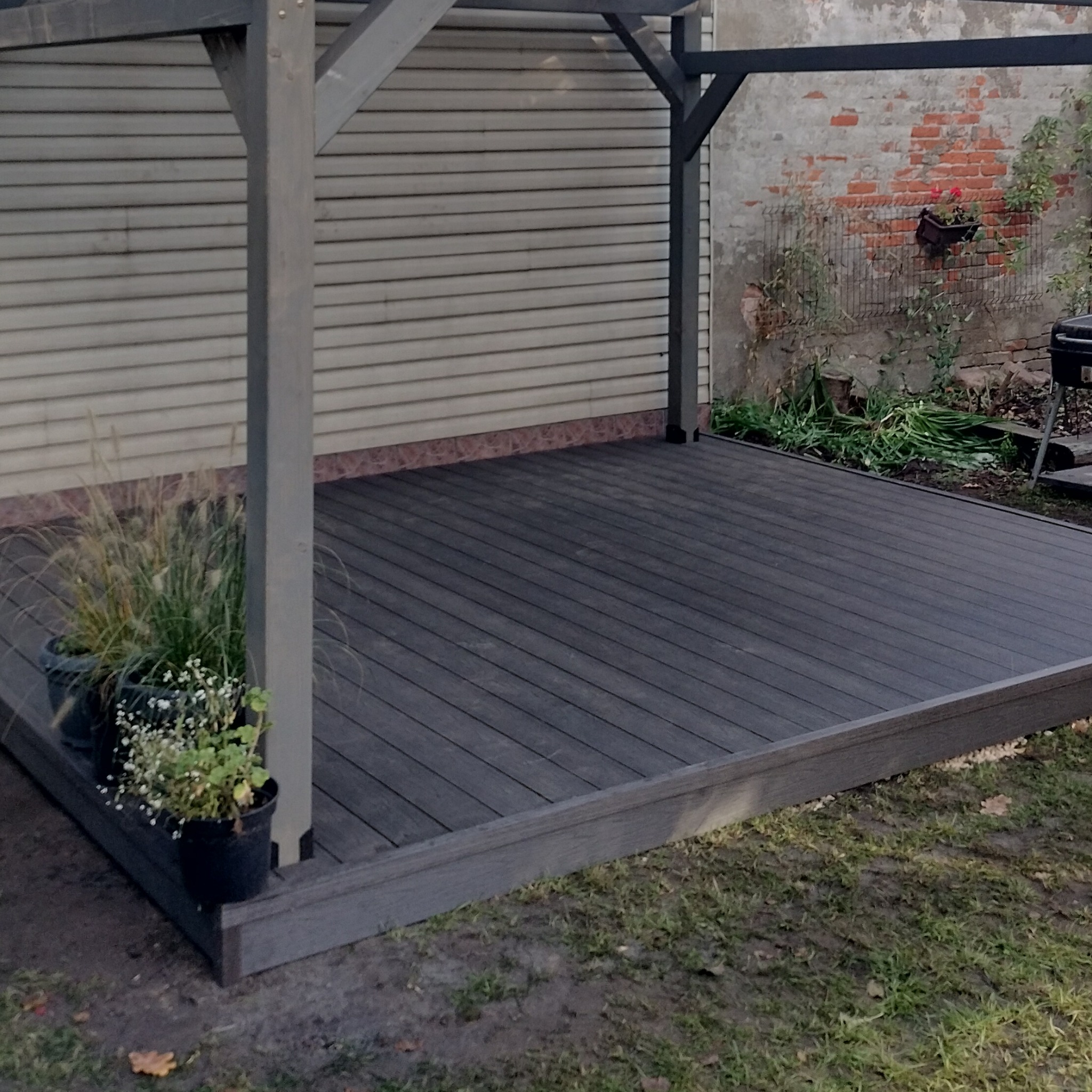 Zadaszony taras z kompozytowych desek w kolorze ciemnego grafitu, otoczony zielenią i doniczkami z roślinami, z widoczną ścianą budynku w tle.