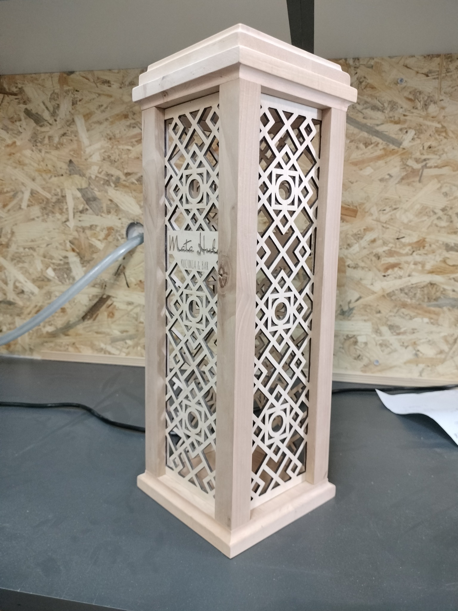 Drewniana kolumna z ażurowym wzorem geometrycznym, jasne drewno, widoczne logo 'Mata Hud Kuchnia & Bar', surowe wykończenie, kabel w tle.