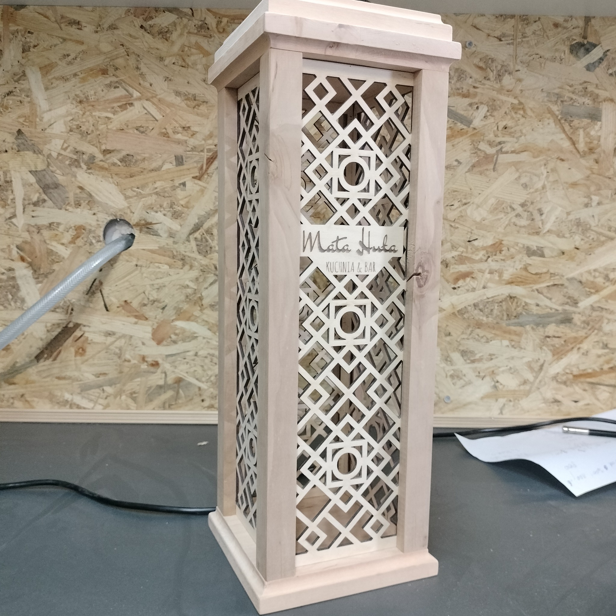 Drewniana lampa z ażurowym wzorem i logo 'Mata Huka KUCHNIA & BAR' na tle płyty OSB, widoczny kabel zasilający oraz notatki.