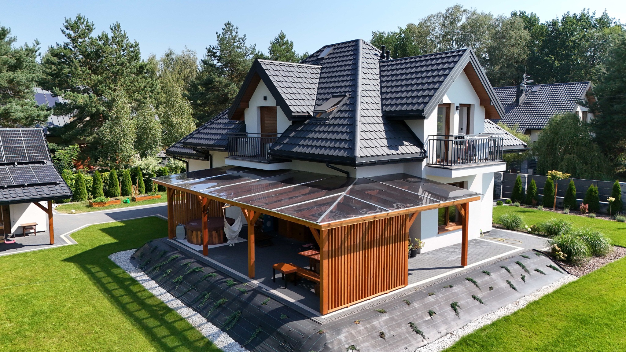 Nowoczesna pergola z przezroczystym zadaszeniem, przylegająca do domu z balkonami, otoczona zadbanym trawnikiem i zielenią, widok z góry.