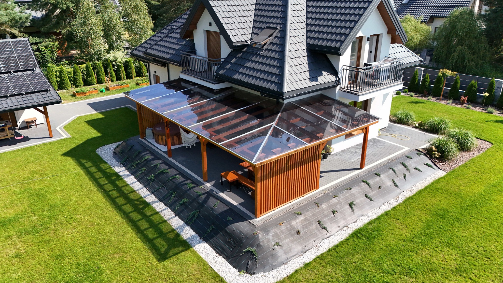Nowoczesna pergola z przezroczystym zadaszeniem przylegająca do domu, widok z góry, z drewnianą ścianą i stolikiem wewnątrz, na tle zadbanego trawnika i kostki brukowej.