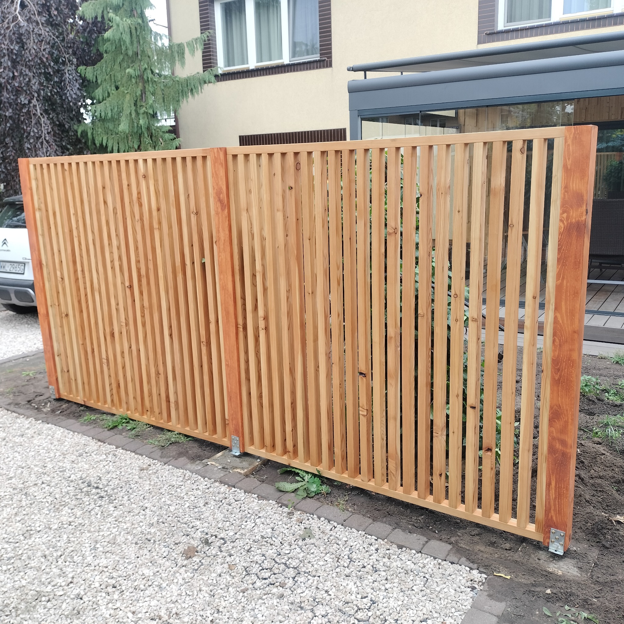 Nowoczesne ogrodzenie z jasnego drewna z pionowymi szczebelkami, widoczne mocowania metalowe, na tle domu z szarą pergolą i żwirowym podłożem.