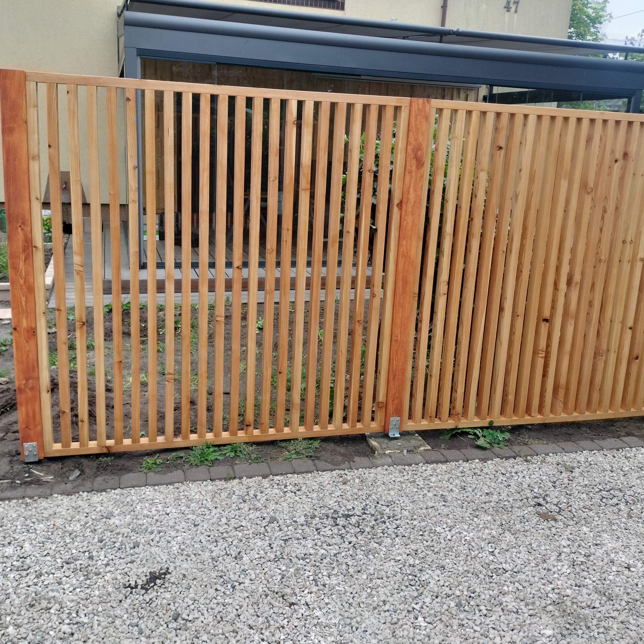 Nowoczesne ogrodzenie z pionowych desek z jasnego drewna, zamontowane na metalowych łącznikach, widoczne na tle szarej nawierzchni z kamieni i fragmentu budynku z pergolą.