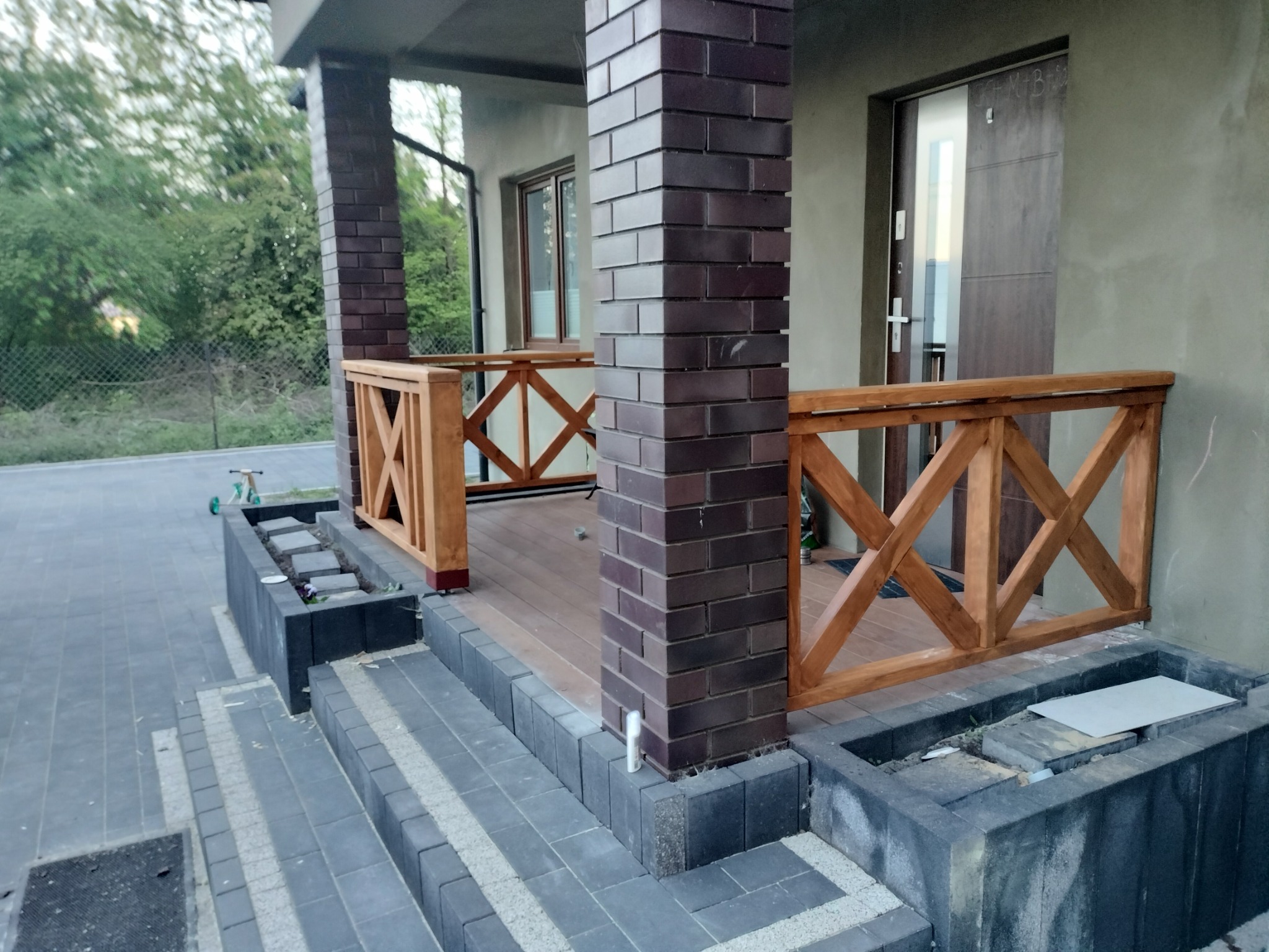 Zadaszony taras z drewnianą balustradą w kształcie krzyży, ceglane filary, schody z szarej kostki brukowej, elementy małej architektury ogrodowej.