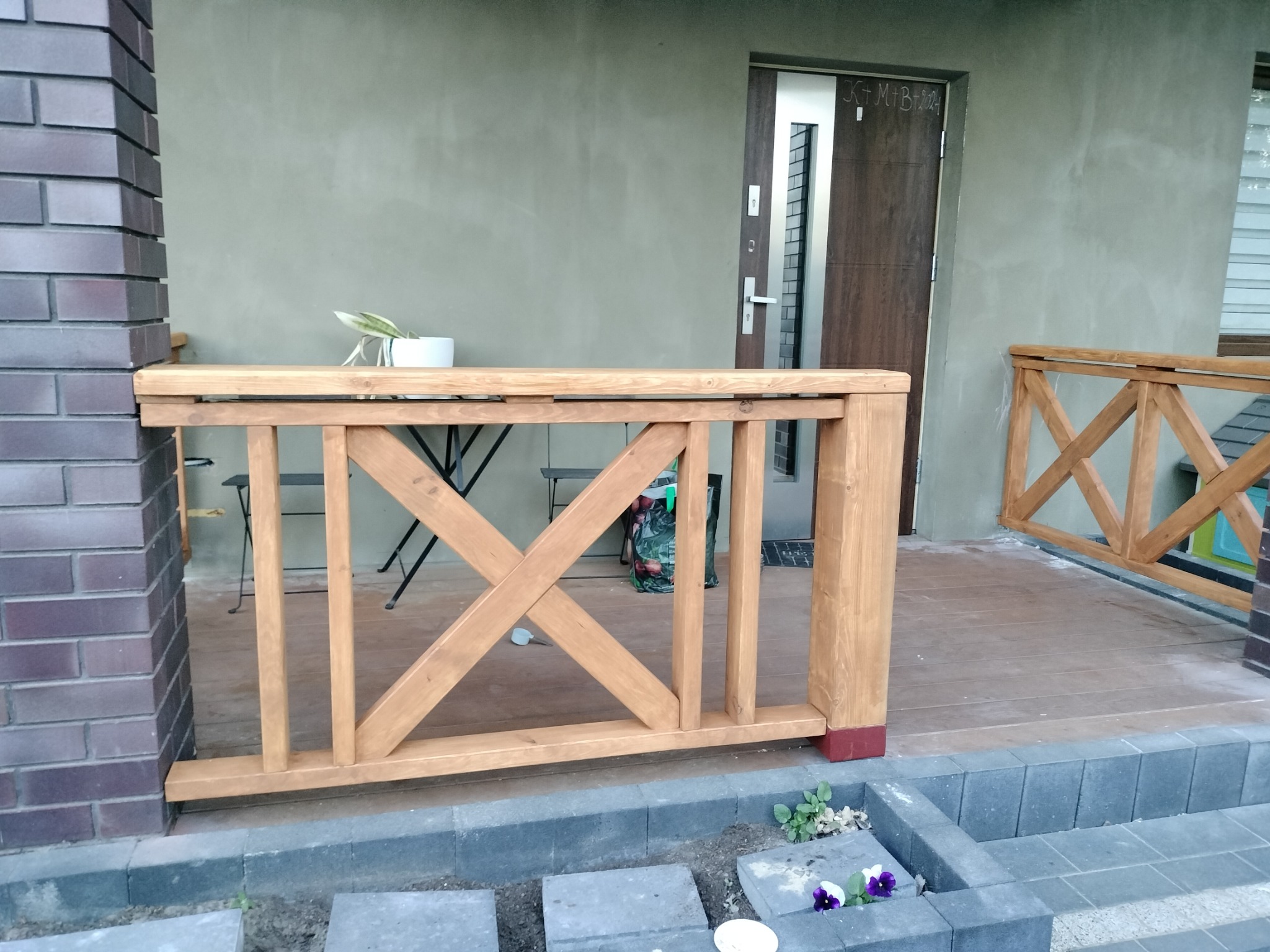 Drewniana balustrada z krzyżowym wzorem, świeżo polakierowana, zamontowana na tarasie w Zagościńcu. Widoczne fragmenty ceglanego filaru i wejścia do domu.