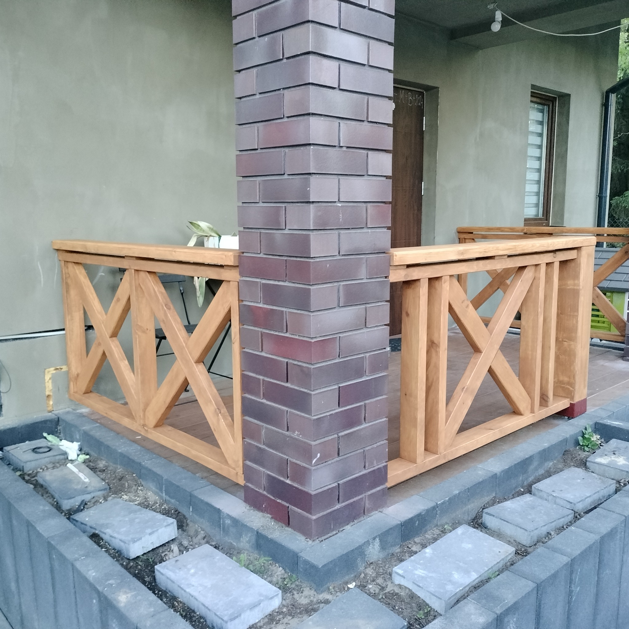 Drewniana balustrada tarasu z krzyżowym wzorem, wsparta na ceglanym filarze, z widocznymi płytami chodnikowymi i fragmentem elewacji budynku.