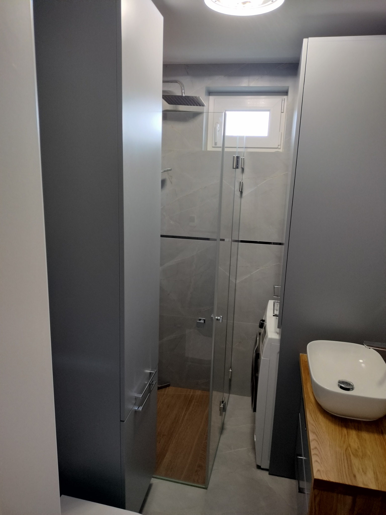 Nowoczesna łazienka z szarą armaturą, kabiną prysznicową ze szklanymi drzwiami, drewnianym blatem pod umywalką i pralką w rogu.