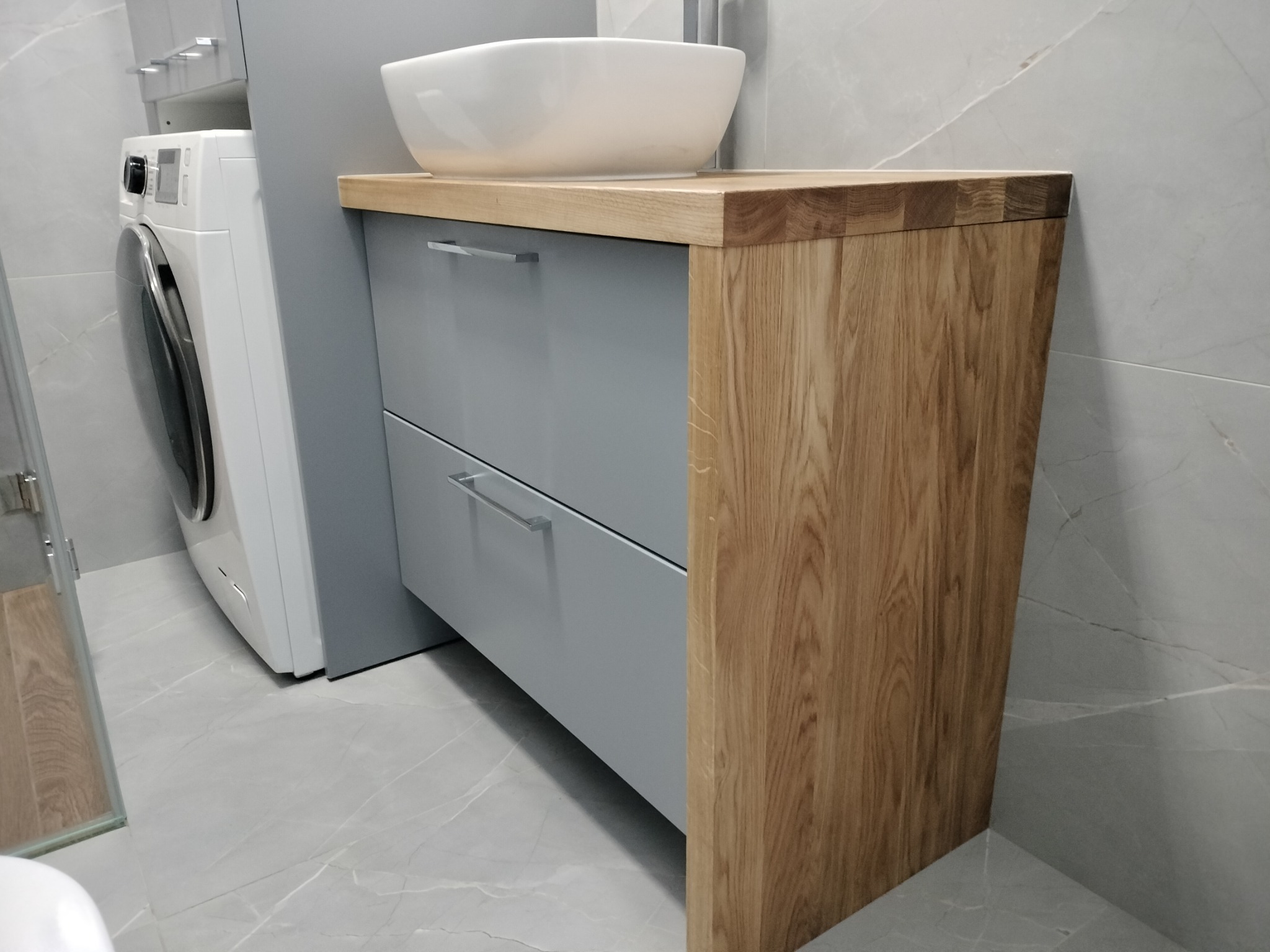 Szafka łazienkowa z drewnianym blatem i umywalką nablatową, szare fronty szuflad, widoczna pralka w tle, płytki podłogowe imitujące marmur.