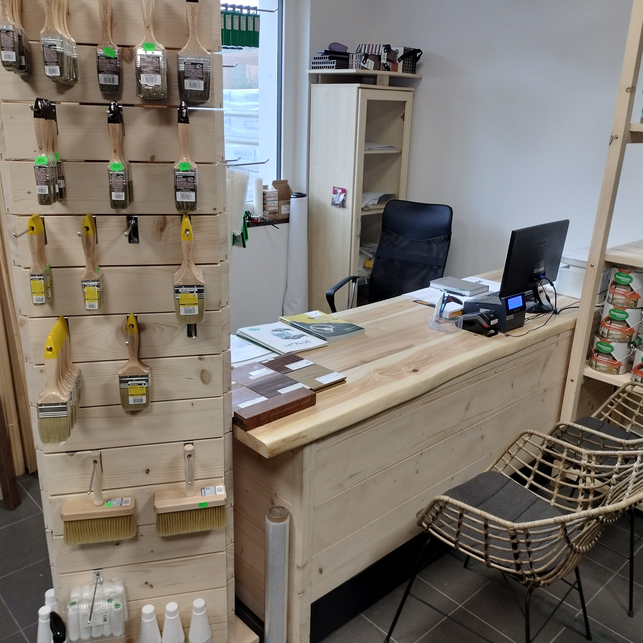 Wnętrze sklepu z materiałami do wykończenia drewna, widoczne pędzle na ekspozytorze, próbki kolorów na biurku z komputerem i kasą fiskalną, oraz krzesła z rattanu.