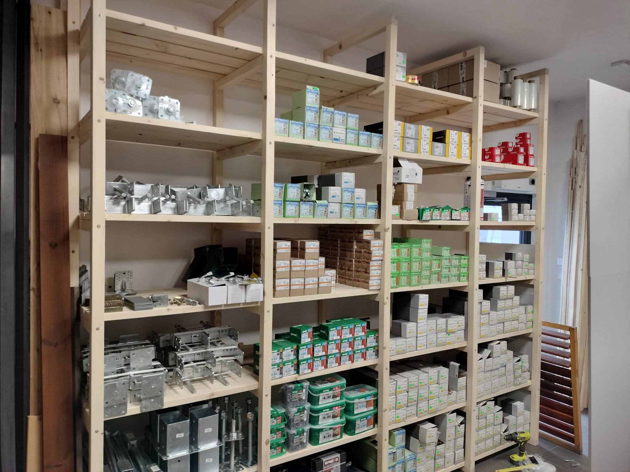 Regał magazynowy z elementami metalowymi i opakowaniami akcesoriów montażowych do konstrukcji drewnianych, widoczne kątowniki, mocowania słupów i wkręty.