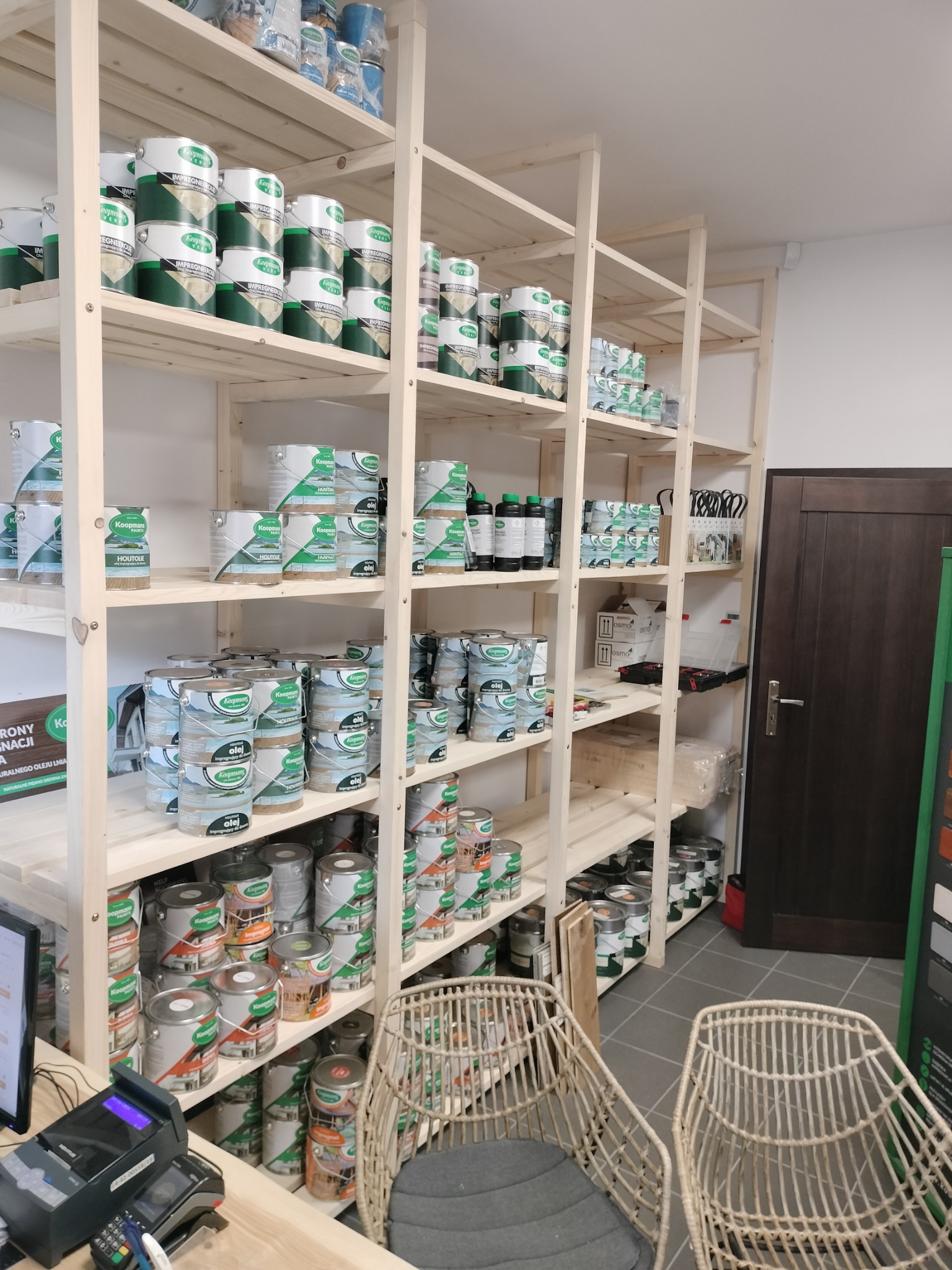 Półki regału w sklepie z farbami i impregnatami do drewna, widoczne puszki z produktami Koopmans i Osmo, krzesła z wikliny w tle.