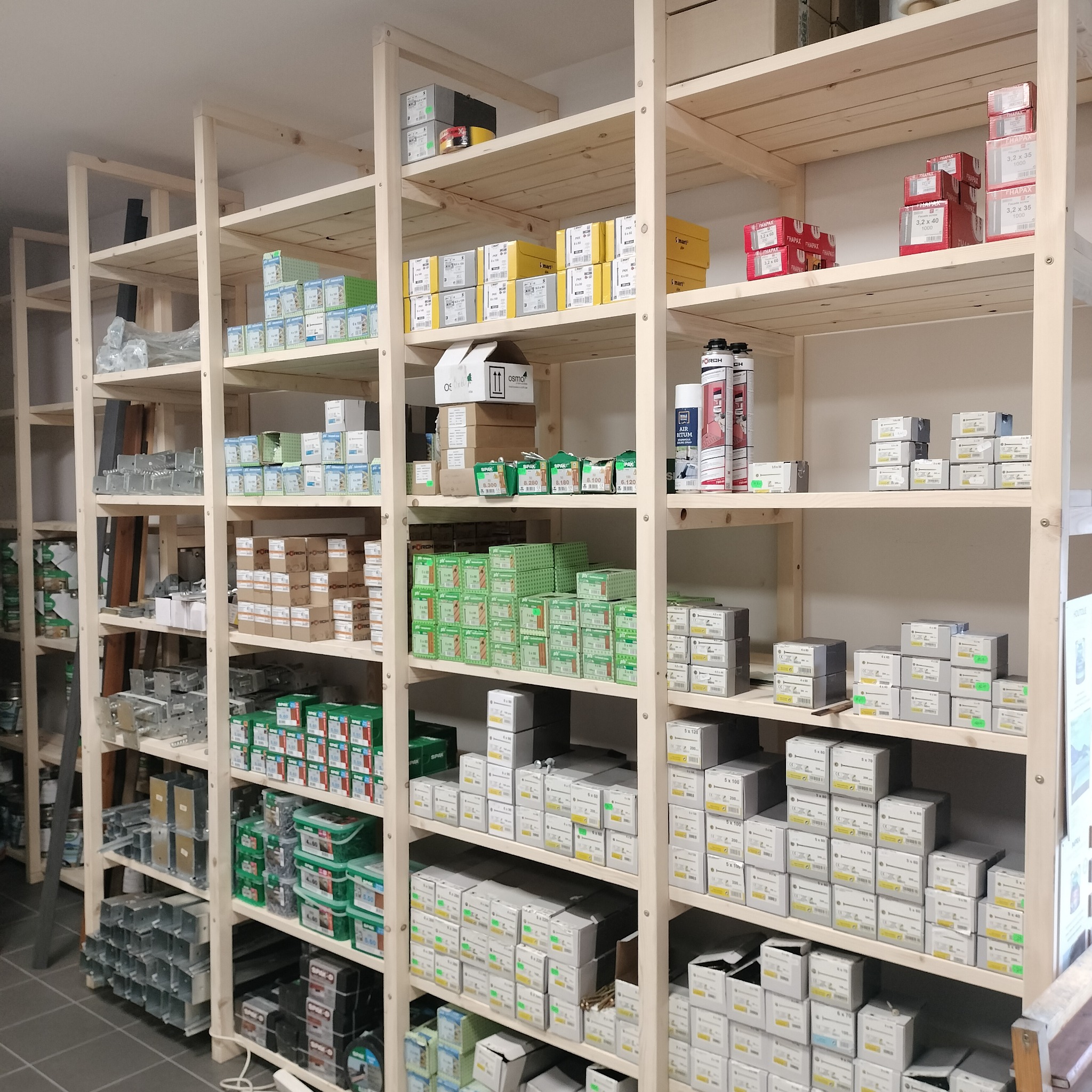 Półki z jasnego drewna wypełnione różnymi materiałami budowlanymi, głównie opakowaniami wkrętów i śrub różnych rozmiarów, oraz puszkami z pianką montażową.