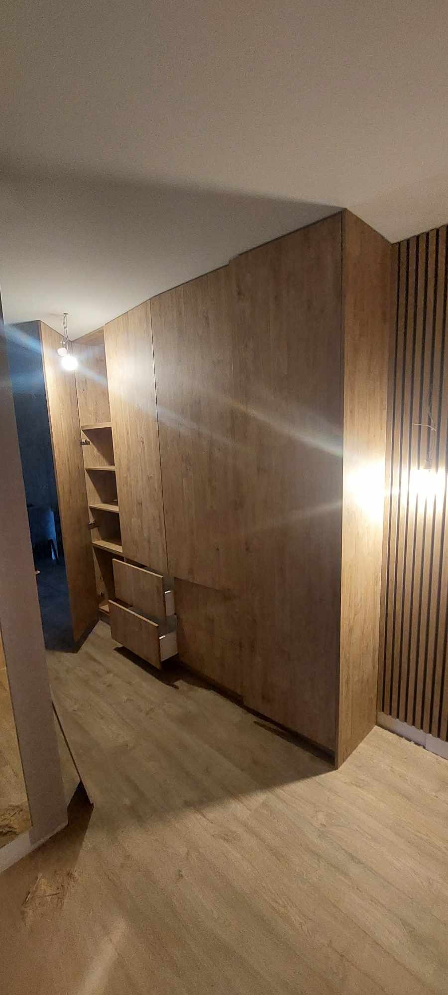 Wnętrze z zabudową meblową z okładziną imitującą drewno, widoczne otwarte półki i wysunięte szuflady, na ścianie panele dekoracyjne.