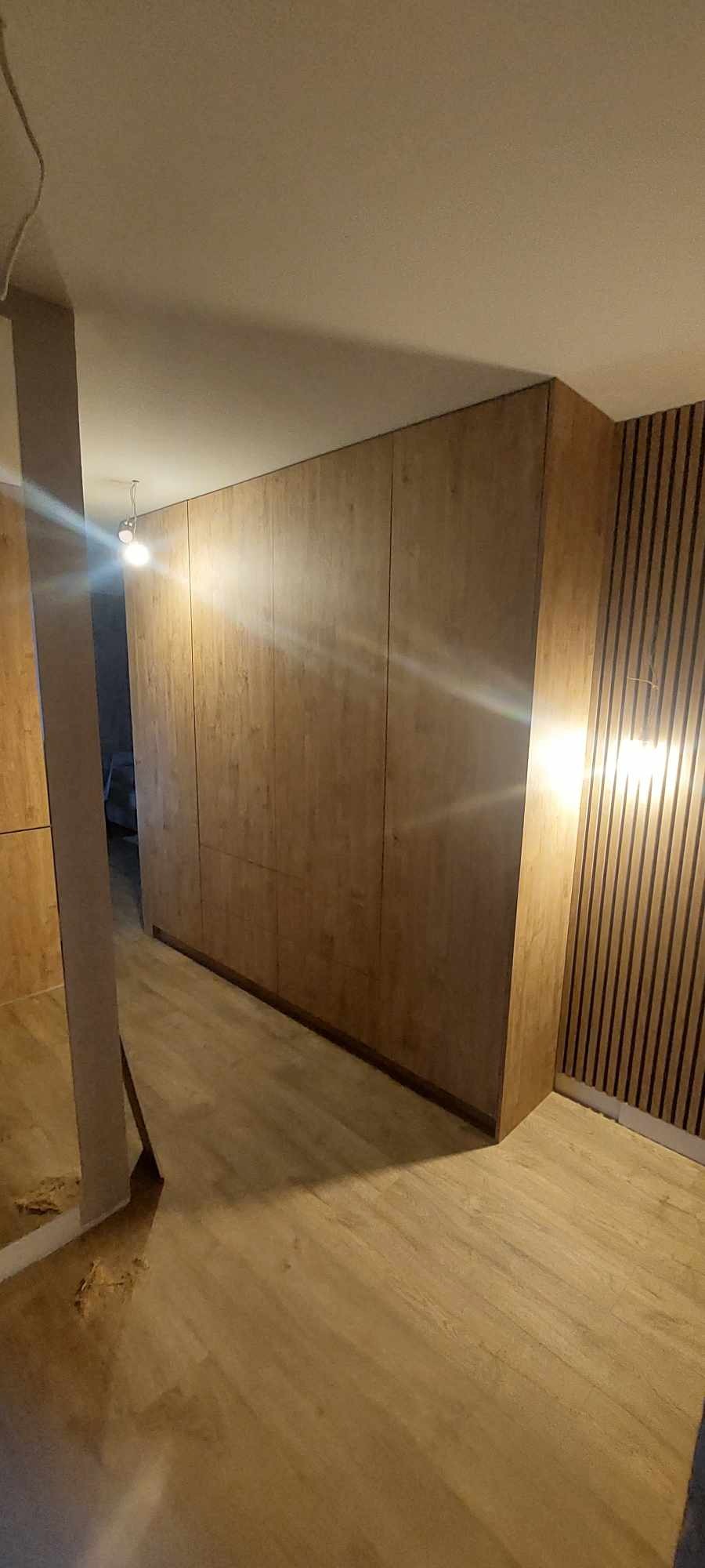 Wnętrze z zabudową szafy z drewnopodobnej płyty, panele ścienne z pionowymi listwami, podłoga z paneli, sufit z oświetleniem.