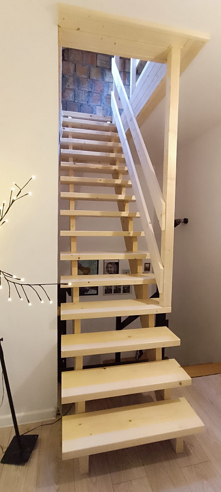 Surowe, drewniane schody z metalowymi wspornikami, prowadzące na strych z ceglaną ścianą, oświetlone dekoracyjną lampą w kształcie gałęzi.