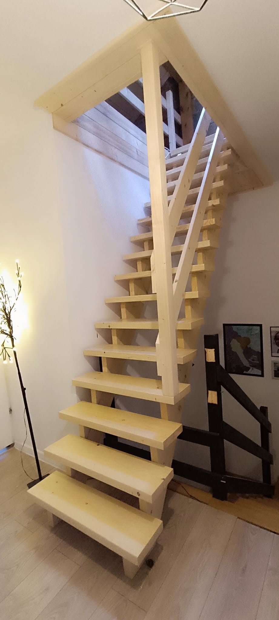 Surowe, jasne drewniane schody wewnątrz domu, z widoczną konstrukcją wsporczą i otwartym prześwitem na górę, oświetlone lampą stojącą z boku.